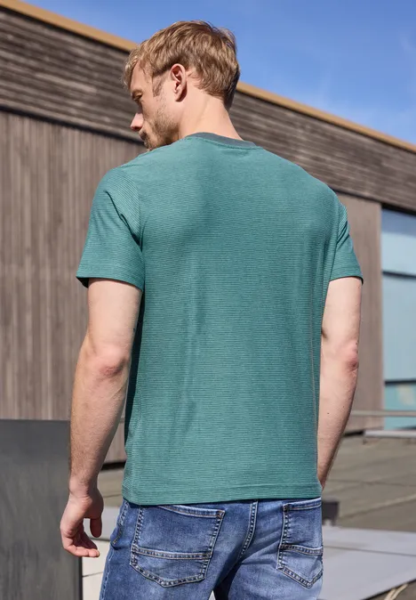 Mann mit grünem T-Shirt und blauer Jeans steht rückwärts vor Holzfassade unter blauem Himmel.