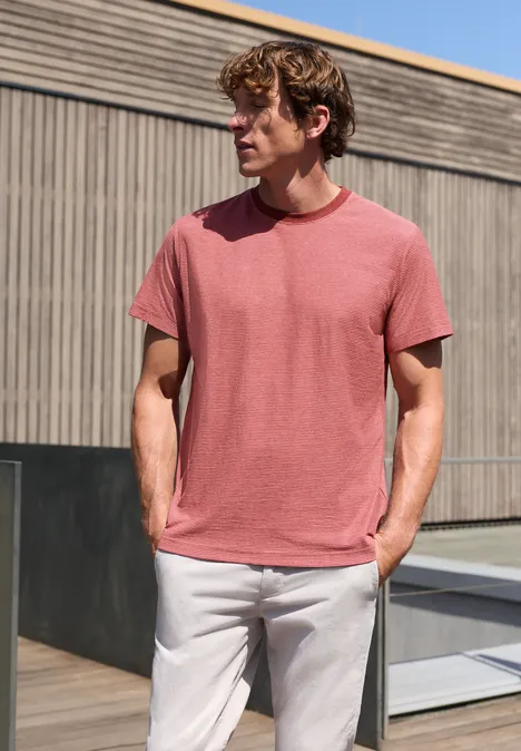 T-Shirt im Yarn Dye Streifen-Look sunrise red Ein Mann in rotem T-Shirt und hellen Hosen steht lässig vor modernem Holzgebäude.