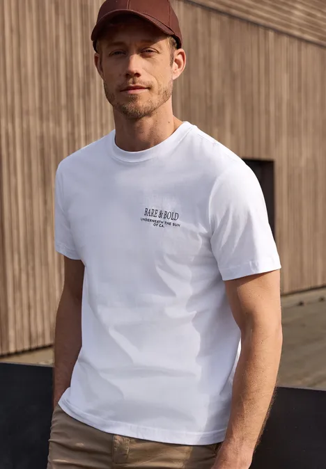 Mann in weißem T-Shirt und roter Kappe steht vor einer Holzfassade.