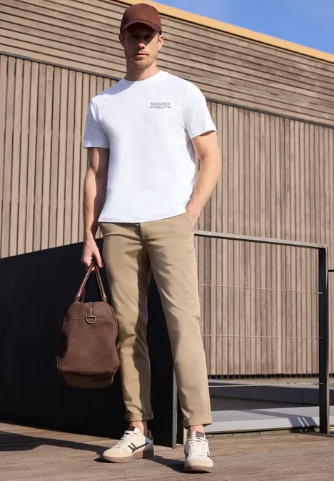 Ein Mann in weißem T-Shirt und beiger Hose hält eine braune Tasche neben moderner Fassade.