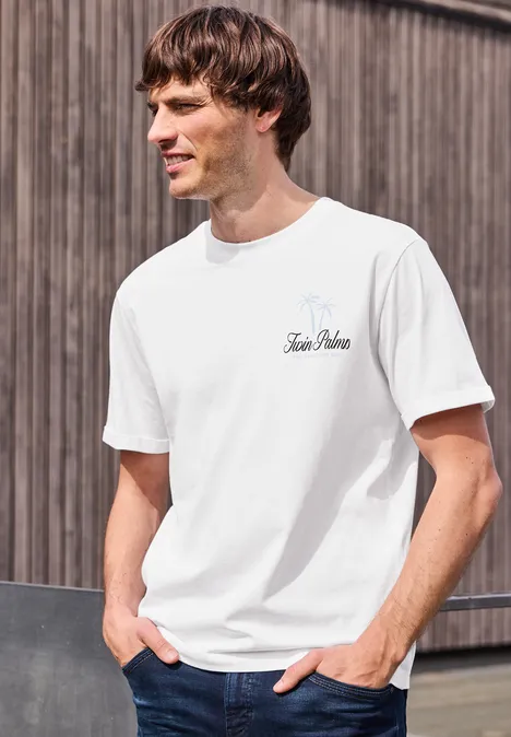 Ein junger Mann lehnt lässig in einem weißen T-Shirt vor einer Holzwand, Hände in den Taschen.