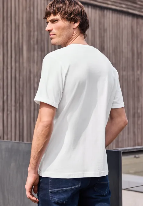 Ein Mann steht seitlich gesehen, trägt ein weißes T-Shirt und blaue Jeans vor einer Holzfassade.