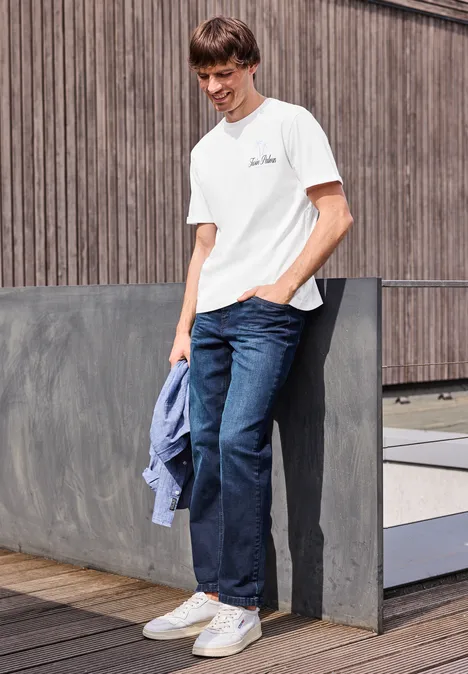 Junger Mann in weißem T-Shirt, Jeans und weißen Sneakern lehnt draußen an Holzwand und hält blaue Jacke.