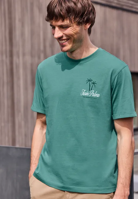 Ein junger Mann trägt ein grünes T-Shirt mit Palme-Logo und Text, mit verschwommenem Gesicht vor einer Holzwand.