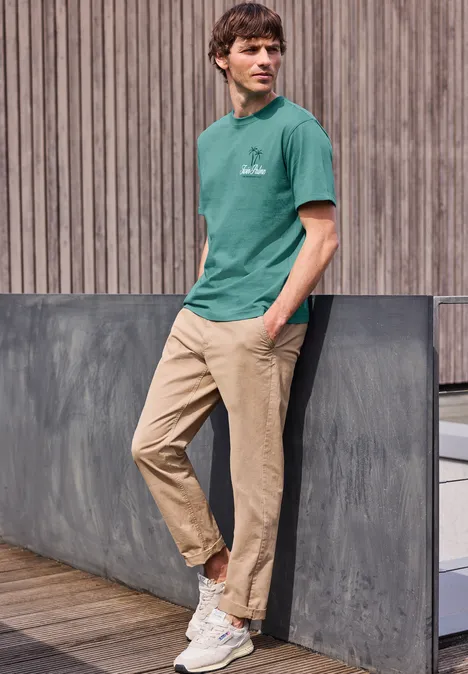 Junger Mann in grünem T-Shirt und beigen Chinos lehnt entspannt an einer Betonmauer vor Holzfassade.