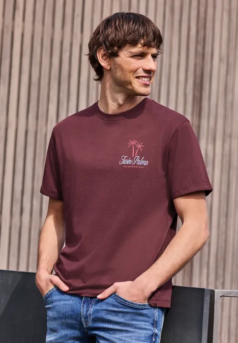 Ein Mann mit dunkelrotem T-Shirt und Jeans steht vor einer Holzfassade und blickt zur Seite.