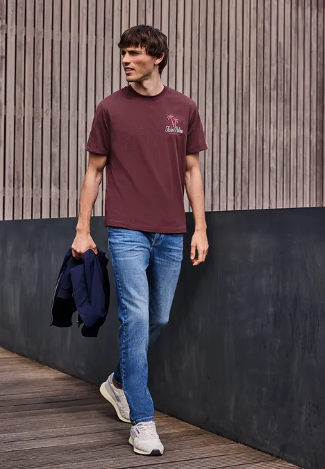 Ein Mann in Jeans und bordeauxrotem T-Shirt geht entlang einer Holzverkleidung und hält eine Jacke.