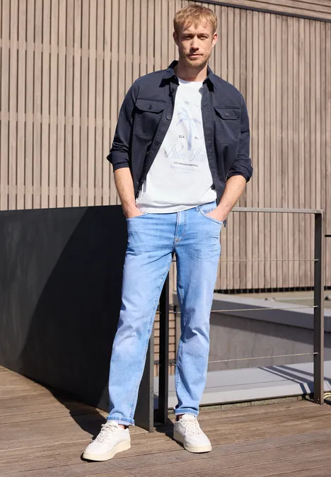 Een jonge man leunt ontspannen tegen een terrasbalustrade, gekleed in jeans en sneakers.
