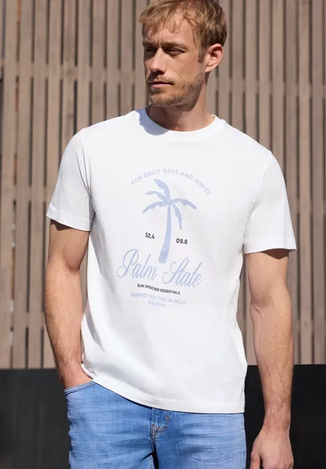 Een man draagt een wit t-shirt met palmboommotief en 'Palm State'-opdruk, met een jeans voor een houten wand.