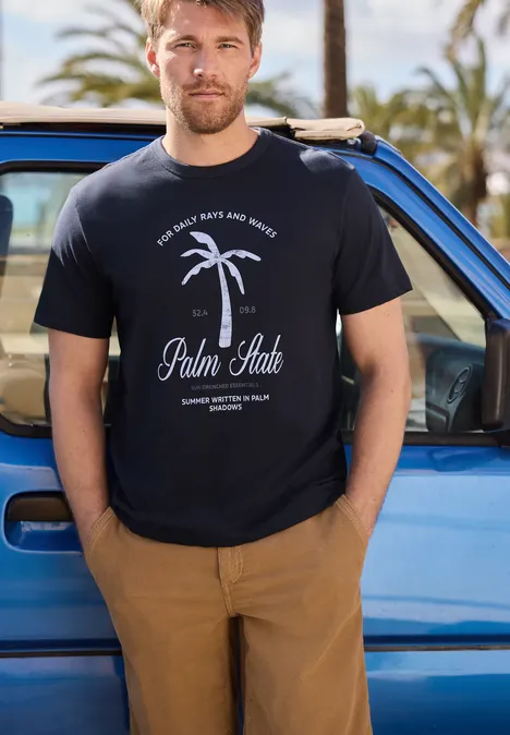Ein Mann steht vor einem blauen Auto und trägt ein T-Shirt mit Palmenmotiv.