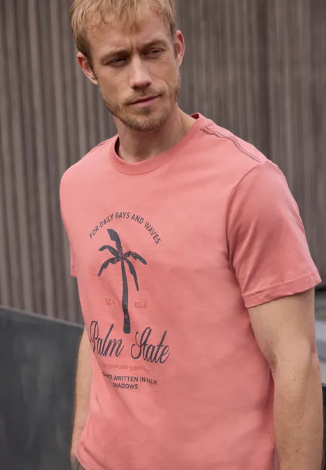 Een man draagt een roze t-shirt met palmbomenprint voor een onscherpe achtergrond.