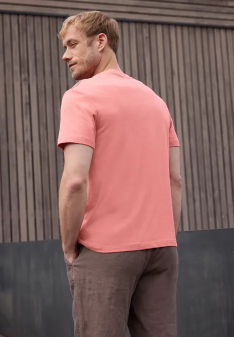 Man in zalmroze T-shirt en bruine broek zijwaarts tegen een houten muur geleund.