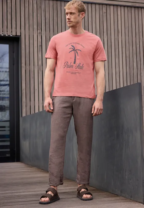 Man in roze T-shirt met palmprint, bruine broek en sandalen op een houten terras.