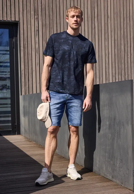 Een man staat op een houten terras, draagt een donker T-shirt, spijkershorts, witte sneakers en houdt een beige pet vast.