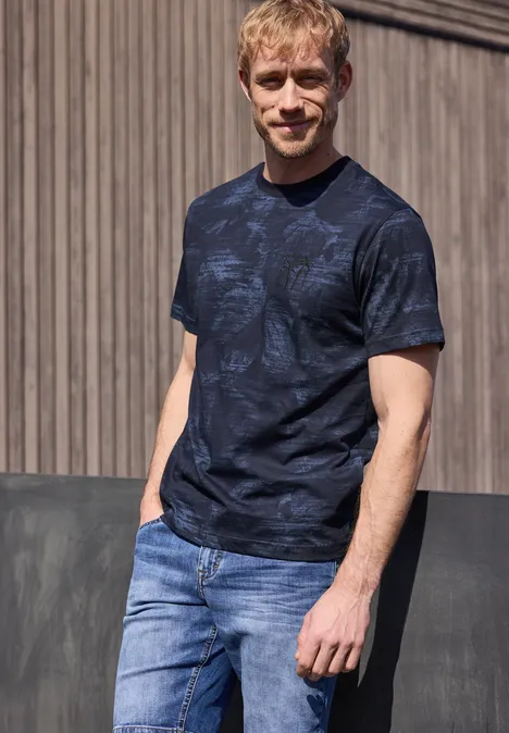 Een man draagt een donkerblauw T-shirt met patroon en een lichtblauwe jeans, leunend tegen een houten wand.