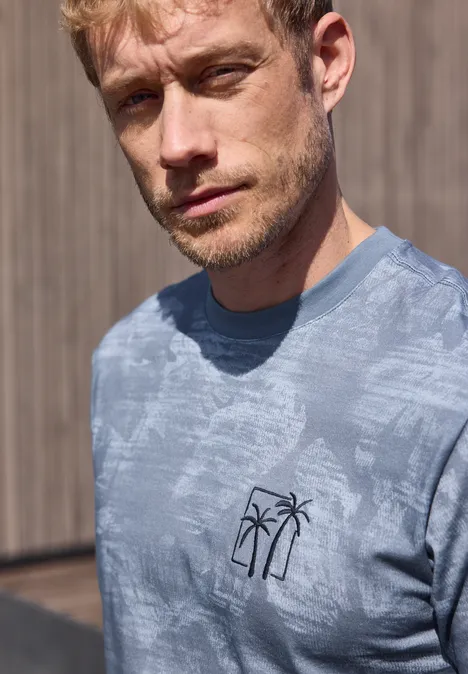 Een blonde man draagt een blauw t-shirt met palmboomlogo, voor een houten wand.