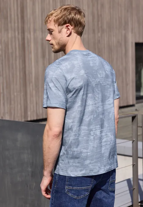 Een man van achteren met een grijs-blauw T-shirt staat voor een houten wand buiten.