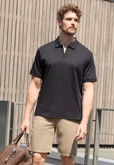 Polo met korte mouwen en ritssluiting dark ink blue Ein Mann mit schwarzem Poloshirt und beigefarbenen Shorts hält eine Tasche vor einer Holzfassade.