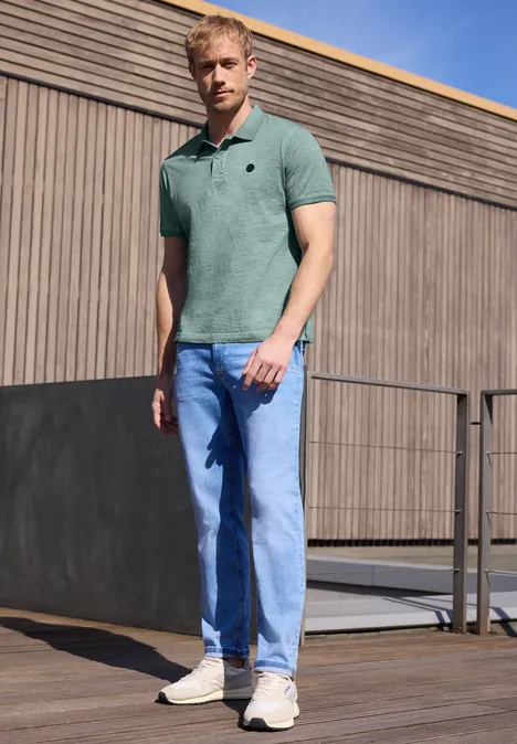 Un homme en polo et jean se tient devant une façade en bois.