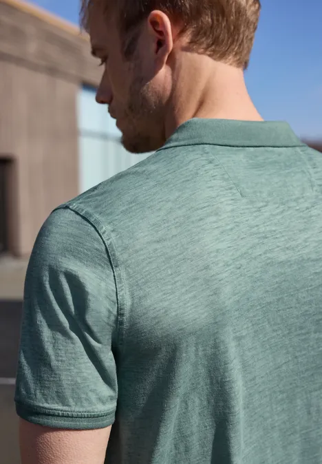 Un homme est vu de dos, portant un polo vert clair, avec un décor extérieur ensoleillé en arrière-plan.