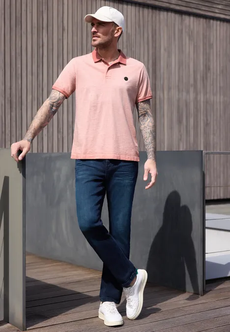 Un homme tatoué porte un polo rose, un jean et des baskets blanches, appuyé contre une façade en bois.