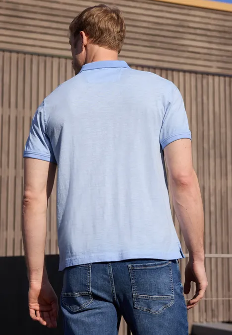 Un homme vu de dos porte un polo bleu clair et un jean devant une façade en bois.