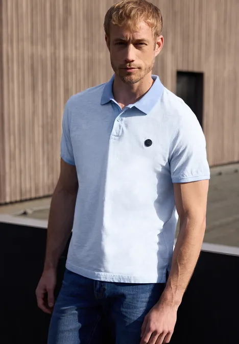 Un homme porte un polo bleu clair et un jean, debout devant une façade en bois en plein air.