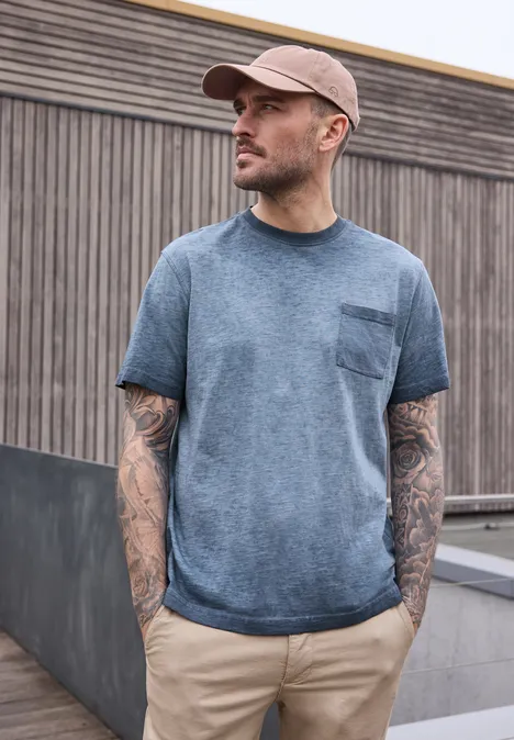 Ein Mann mit Tattoo-ärmeln in blauem T-Shirt und Kappe steht vor einer Holzwand.