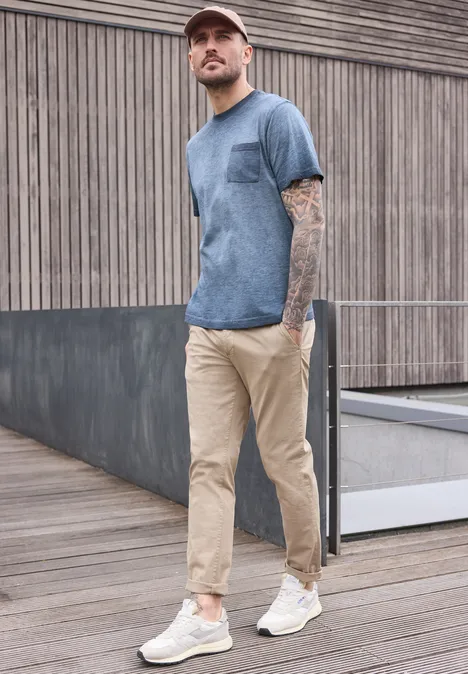 Ein verschwommener Mann in blauem T-Shirt, beiger Chinohose und weißen Sneakern steht entspannt draußen.