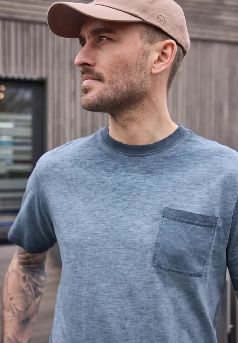 Ein Mann in grauem T-Shirt mit Brusttasche, Baseballcap und sichtbaren Tattoos vor einer Holzfassade.