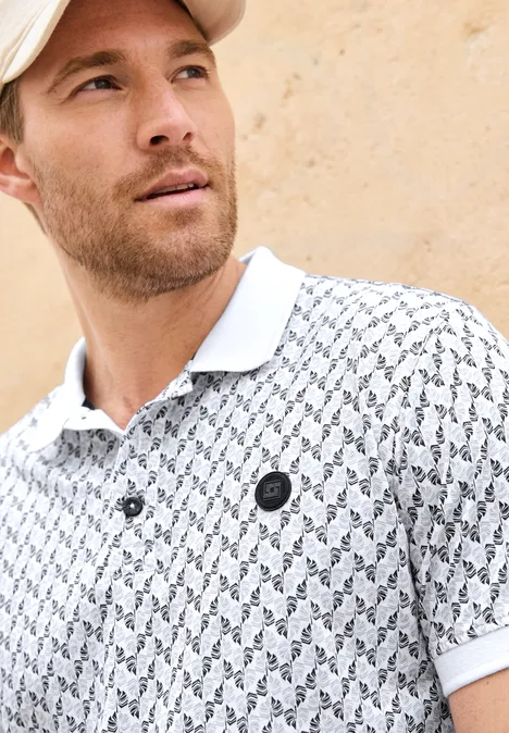 Mann mit Polohemd in Weiß mit kleinem schwarzem Logo und Muster vor beiger Wand.