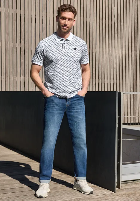 Ein Mann in weiß gemustertem Poloshirt, blauen Jeans und weißen Sneakern steht draußen vor Holzwand.