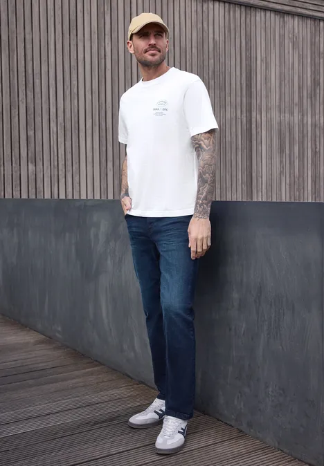 Ein tätowierter Mann steht in weißem T-Shirt, blauen Jeans und weißen Sneakern vor einer Holz- und Stahlwand.