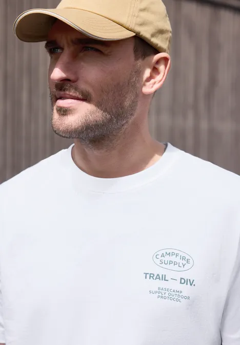 Mann mit Baseballcap und weißem T-Shirt mit Logo vor einer Holzfassade.