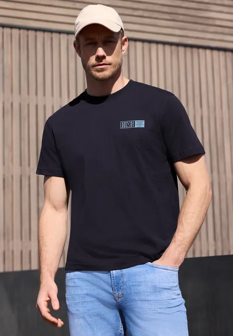 Ein Mann in schwarzem T-Shirt und blauer Jeans steht vor einer Holzwand.