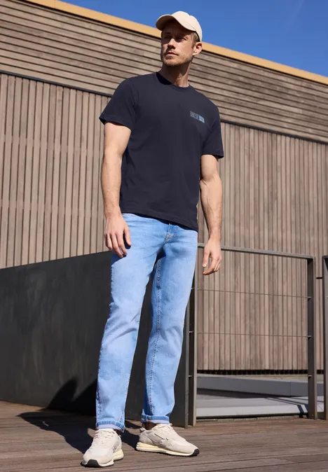 Ein Mann in schwarzem T-Shirt, hellblauen Jeans und weißen Sneakern steht draußen an einer Holzfassade.