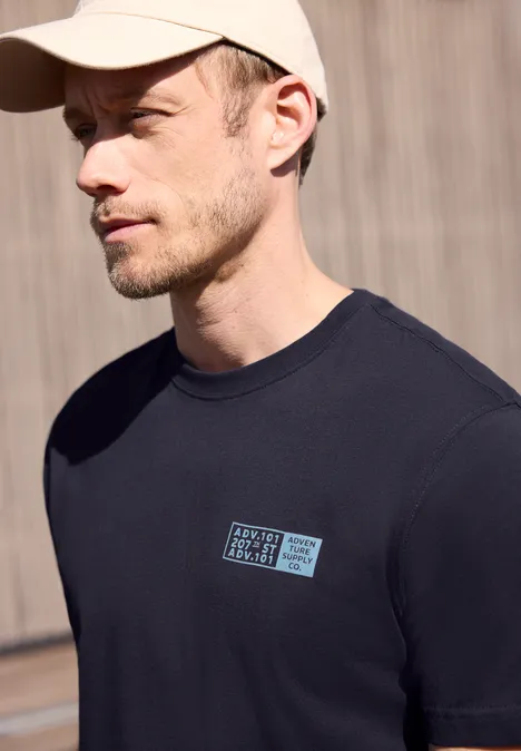 Seitliche Ansicht eines Mannes mit schwarzem T-Shirt und Kappe vor einer Holzwand, Gesicht unkenntlich gemacht.