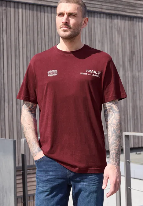 Mann steht lässig draußen, trägt rotes T-Shirt mit Aufdruck, Jeans und hat tätowierte Arme.