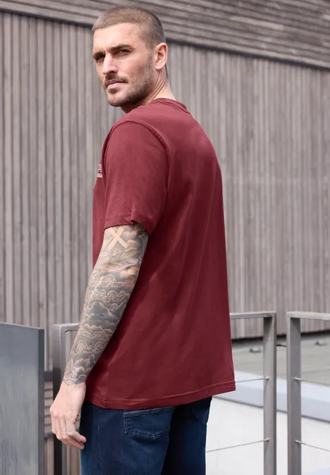 Junger Mann im roten T-Shirt mit Tattoos am Arm steht seitlich vor hölzerner Fassade.