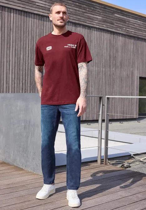 Ein Mann mit rotem T-Shirt, blauer Jeans und weißen Sneakern steht auf einer Holzterrasse vor einem Gebäude.