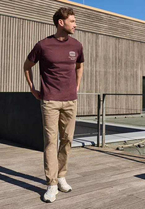 Ein Mann steht auf einer Holzterrasse, trägt ein bordeauxfarbenes T-Shirt, eine beige Hose und weiße Sneaker.