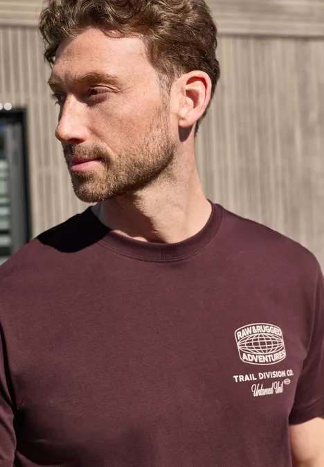 Ein junger Mann trägt ein dunkelrotes T-Shirt mit weißem Print und steht vor einer Holzvertäfelung.