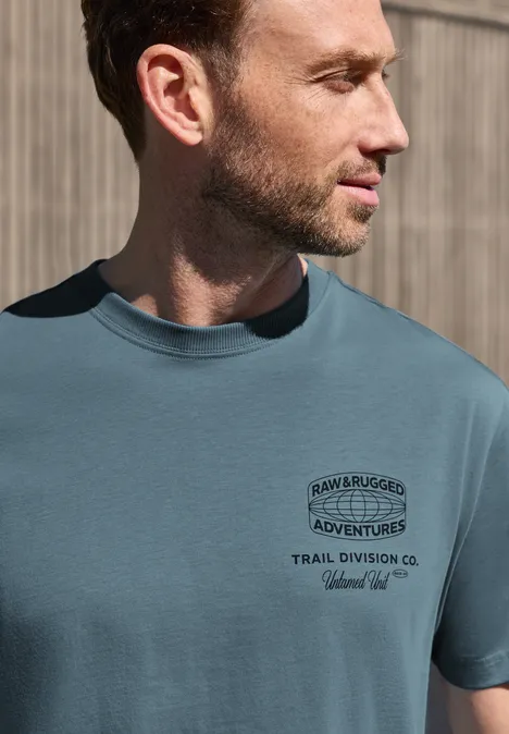 Ein seitliches Porträt eines Mannes, der ein türkisfarbenes T-Shirt mit Abenteuer-Print trägt.