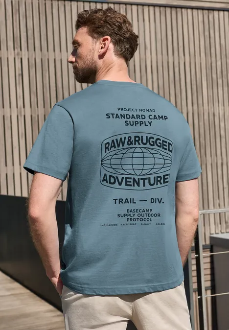 Mann mit türkisfarbenem T-Shirt und beigen Shorts lehnt an Holzlattenzaun, Rückansicht.