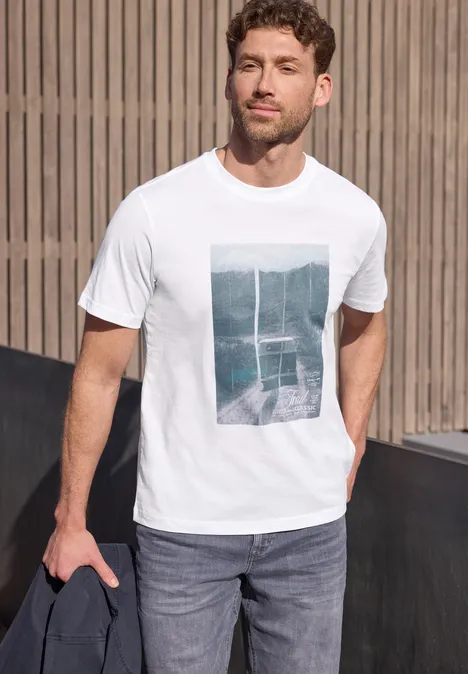 Mann in weißem T-Shirt mit Grafik, blauen Jeans und dunkler Jacke in der Hand vor Holzfassade stehend.