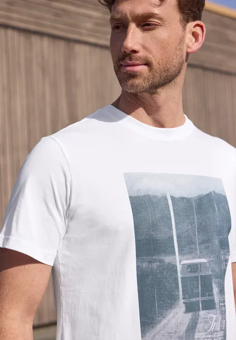 Ein bärtiger Mann trägt ein weißes T-Shirt mit Bergblick und Seilbahn vor Holzwand.