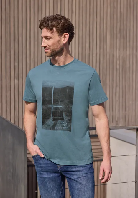 Ein Mann in türkisfarbenem T-Shirt mit Print und Jeans steht lässig vor einer holzverkleideten Wand.