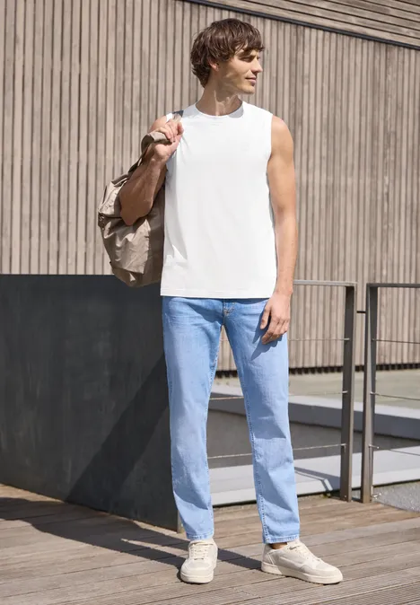 Ein junger Mann vor einer Holzfassade, trägt weißes ärmelloses Shirt, helle Jeans, Sneakers und Umhängetasche.