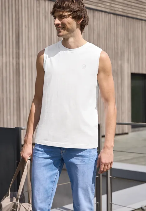 Een man in een wit mouwloos shirt en lichte spijkerbroek op een houten terras, met een beige tas.