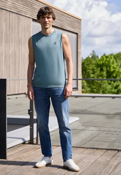 Een man in jeans en mouwloos shirt staat op een houten terras voor een modern gebouw en groen.